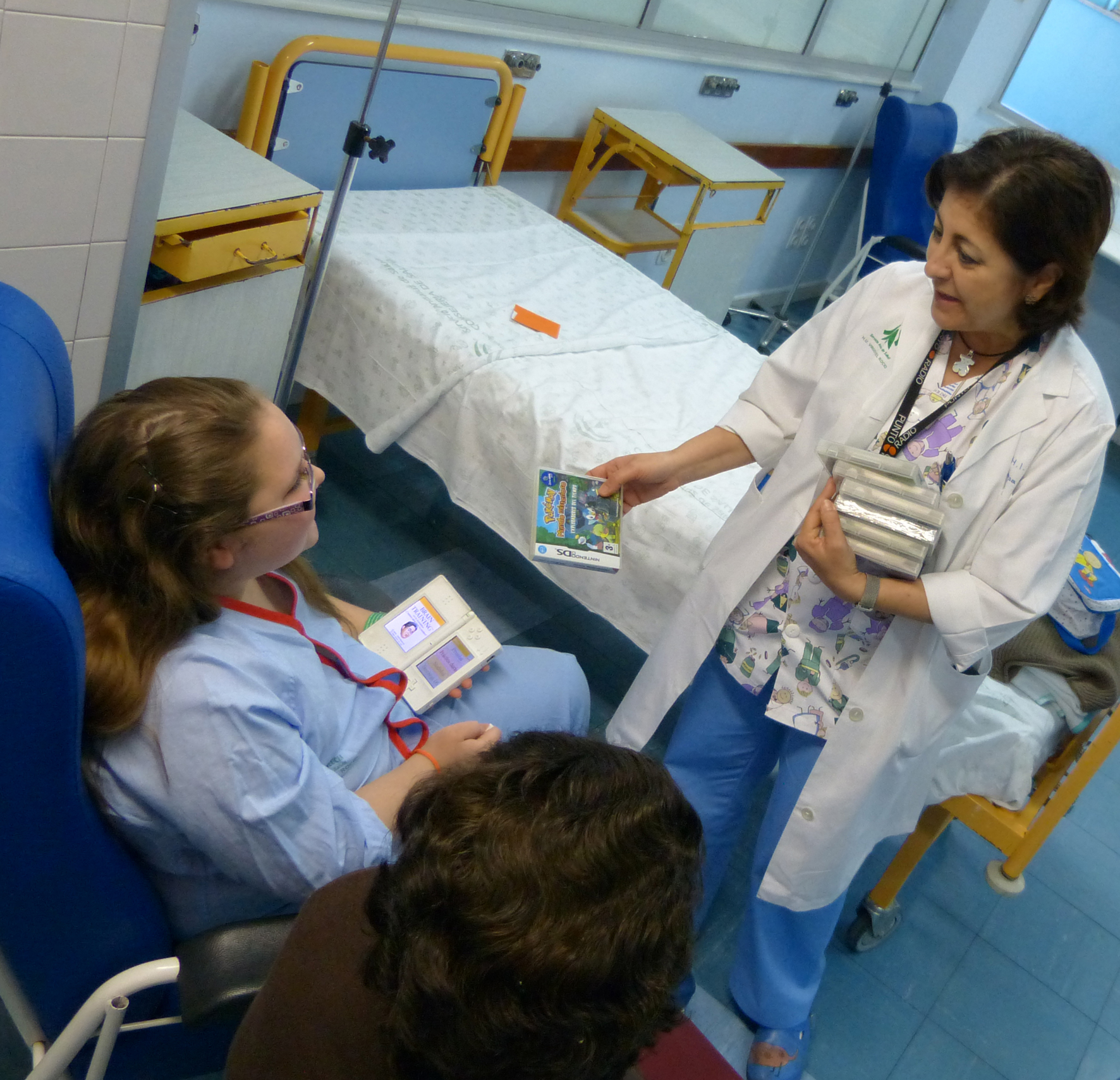 Una niña de la planta de Cirugía Pediátrica recibe de una enfermera un juego para su consola de 'Juegaterapia'